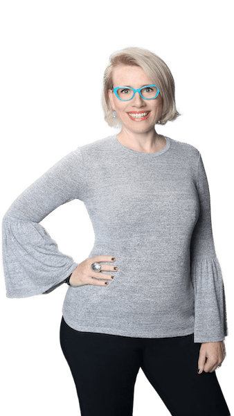 Dr. Shannon Coates The Vocal Instrument 101 Follow Up Teacher Training Practicum creator, Dr. Shannon Coates, wearing teal-framed glasses, a grey shirt with wing arms, and black pants, strikes a jaunty pose with her right hand on her hip. She smiles openly at the camera.
