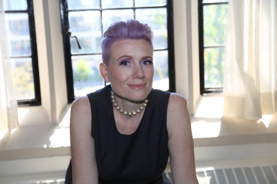 Shannon Coates, speaker for masterclasses and workshops, dressed in a black tank top dress and wearing a short necklace with large, luminescent beads, Dr. Shannon Coates leans forward into the camera. Her hair is short and purple, and her eyes look into the camera while her head tilts upward to the right.