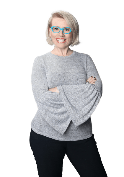 Dr. Shannon Coates: The Vocal Instrument 101 LIVE The Vocal Instrument 101 LIVE creator, Dr. Shannon Coates, wearing teal-framed glasses, a grey shirt with wing arms, and black pants, strikes a jaunty pose with her arms crossed in front of her body. She smiles widely at the camera.