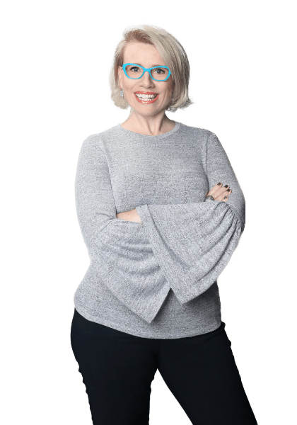 The Vocal Instrument 101 LIVE creator, Dr. Shannon Coates, wearing teal-framed glasses, a grey shirt with wing arms, and black pants, strikes a jaunty pose with her arms crossed in front of her body. She smiles widely at the camera.